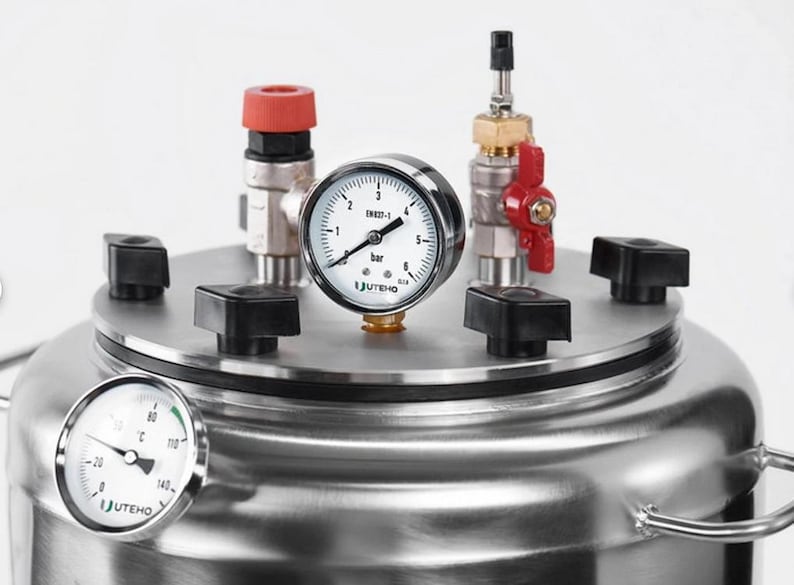 May include: A stainless steel pressure vessel featuring a top lid secured with four black knobs. The lid includes a pressure gauge labelled bar, a red-handled valve, and a red-capped fitting. A temperature gauge is mounted on the side of the vessel.