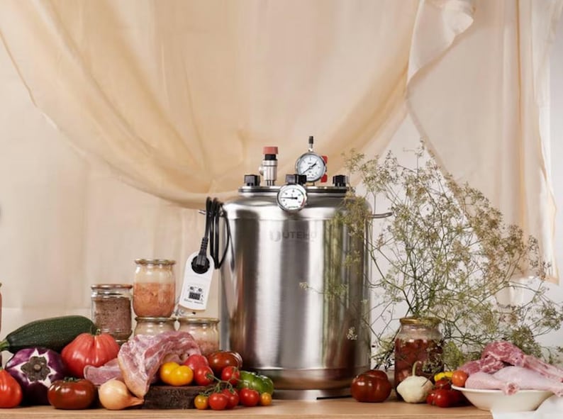 May include: A stainless steel pressure canner with a digital temperature controller, two analogue pressure gauges, and a red valve on top. The unit is surrounded by fresh produce including tomatoes, onions, aubergine, courgette, and raw meat in jars.