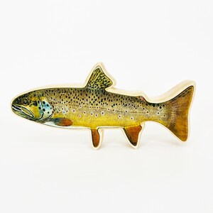 May include: A wooden cutout of a brown trout fish with realistic details. The fish is painted with brown, yellow, and black markings. The cutout is a decorative item for a home or office.