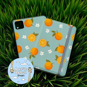 Peut inclure: Une coque de téléphone bleu clair et un journal assorti avec un motif d'oranges, de fleurs blanches et de feuilles vertes. Le design comprend des oranges entières et tranchées. Un autocollant rond avec des girafes et le texte "A Rise Above Designs" est également visible.