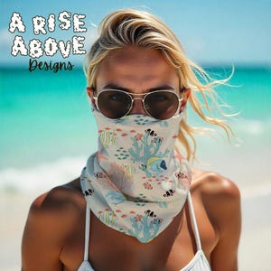May include: A cream-colored face covering with a colorful coral reef and fish pattern. The design includes yellow, blue, and red fish, along with blue coral and red sea plants. The text "A Rise Above Designs" is at the top.