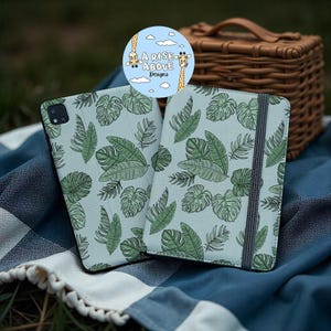 Può includere: Due custodie per tablet con sfondo azzurro e motivo a foglie verdi. Una custodia è aperta, rivelando l'interno. Le custodie sono su una coperta a scacchi blu e bianca. Un logo rotondo con giraffe e il testo "A Rise Above Designs" è visibile.