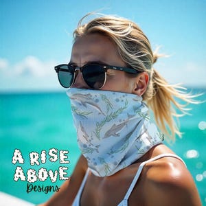 Puede incluir: Una mascarilla azul claro con un estampado de delfines y algas marinas. El texto "A Rise Above Designs" se muestra en blanco, con una fuente estilizada. La persona lleva gafas de sol.
