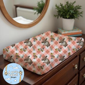 Puede incluir: Una funda de cambiador de color rosa y blanco a cuadros con un patrón floral y lindas vacas de dibujos animados. La funda de cambiador está sobre una mesa de cambio de madera.