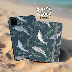 Pode incluir: Uma capa de telefone e um caderno com um design de golfinhos a condizer. O fundo azul marinho apresenta golfinhos brancos, algas verdes e bolhas. O texto "A Rise Above Designs" está no canto superior direito.