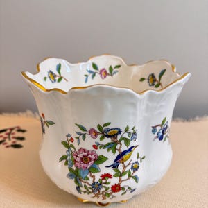 May include: A white porcelain bowl with a scalloped rim and a floral design. The bowl features a bird perched on a branch with flowers and leaves in shades of pink, blue, yellow, and green. The bowl has a gold trim around the rim and base.