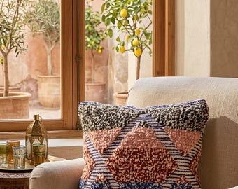 May include: A square throw pillow featuring a textured, tufted geometric pattern in shades of black, dusty pink, rust red, blue, and mustard yellow, set against a cream-colored fabric background with thin horizontal stripes throughout the design.