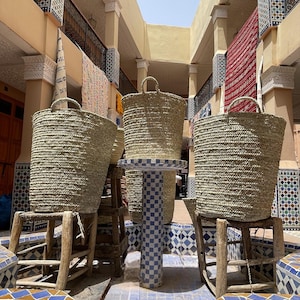May include: Large, woven straw baskets with handles sit on wooden stools. The baskets are cylindrical and a natural, light brown colour. The scene is set in an outdoor market with colourful tilework and architectural details.