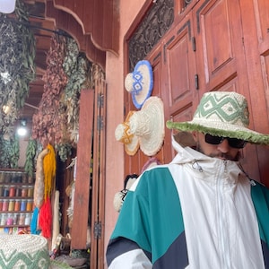 May include: A selection of straw hats with geometric designs and green edging. The hats are displayed on a wooden door. A person wearing a green, white, and black jacket and sunglasses is in the foreground. The background features dried herbs and spices.