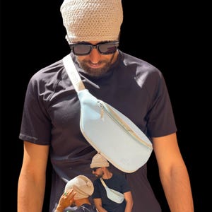 May include: A light blue bum bag with a white strap and a gold zip. The bag is worn across the body. The person in the image is wearing a black t-shirt, sunglasses, and a light beige knitted beanie.