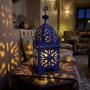 May include: A blue metal lantern with intricate geometric cutouts that cast warm, glowing light patterns onto the surrounding wall. The lantern features a domed top with a decorative finial and a square base, emitting a soft, golden illumination.