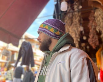 May include: A purple knit beanie with yellow and green geometric patterns. The hat is worn by a person wearing a green hoodie and a white jacket with black stripes. The background is a blurred outdoor market scene.
