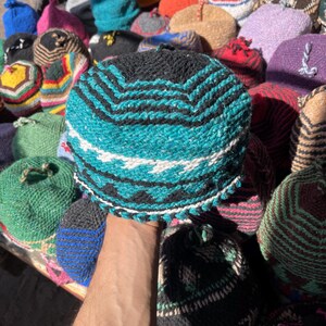 May include: A collection of handmade knit hats in various colors and patterns. The foreground hat is teal, black, and white with a chevron design. Other hats include blue, pink, yellow, and brown, showcasing diverse textures and styles.