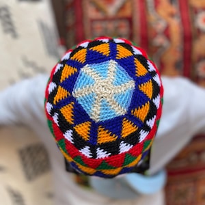 May include: A colorful, patterned knit cap featuring a geometric design in blue, yellow, black, red, green, and white. The cap is viewed from above, showcasing the intricate pattern and vibrant colors. The cap is a traditional style.