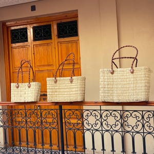 May include: Three rectangular woven straw tote bags with brown leather handles and accents. The bags are displayed on a black wrought iron railing, with a wooden door and tan wall in the background. The bags are of varying sizes.