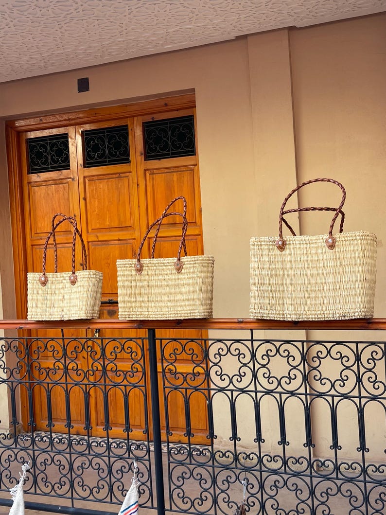 May include: Three rectangular woven straw tote bags with brown leather handles and accents. The bags are displayed on a black wrought iron railing, with a wooden door and tan wall in the background. The bags are of varying sizes.