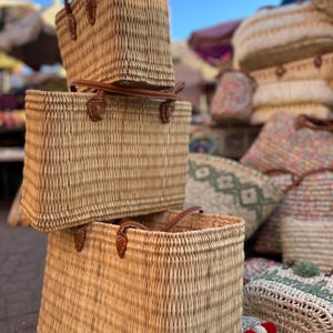 May include: Woven straw tote bags with brown leather handles. The bags are rectangular with a basket weave design. Other bags with floral and geometric patterns are in the background.