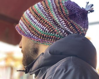 May include: A handmade crochet beanie in a mix of colors including purple, orange, green, brown, and white. The hat has a textured pattern and a small tassel detail at the top. The beanie is worn by a person wearing a black jacket.