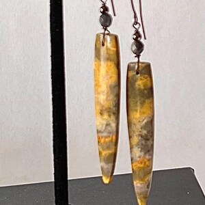 以下が含まれることがあります： 黄色、グレー、茶色の模様が入った、長方形のイヤリングのペア。 イヤリングは濃い色の金属製のフックから吊り下げられており、本体の上には小さな灰色のビーズが付いています。 イヤリングは黒いスタンドに展示されています。