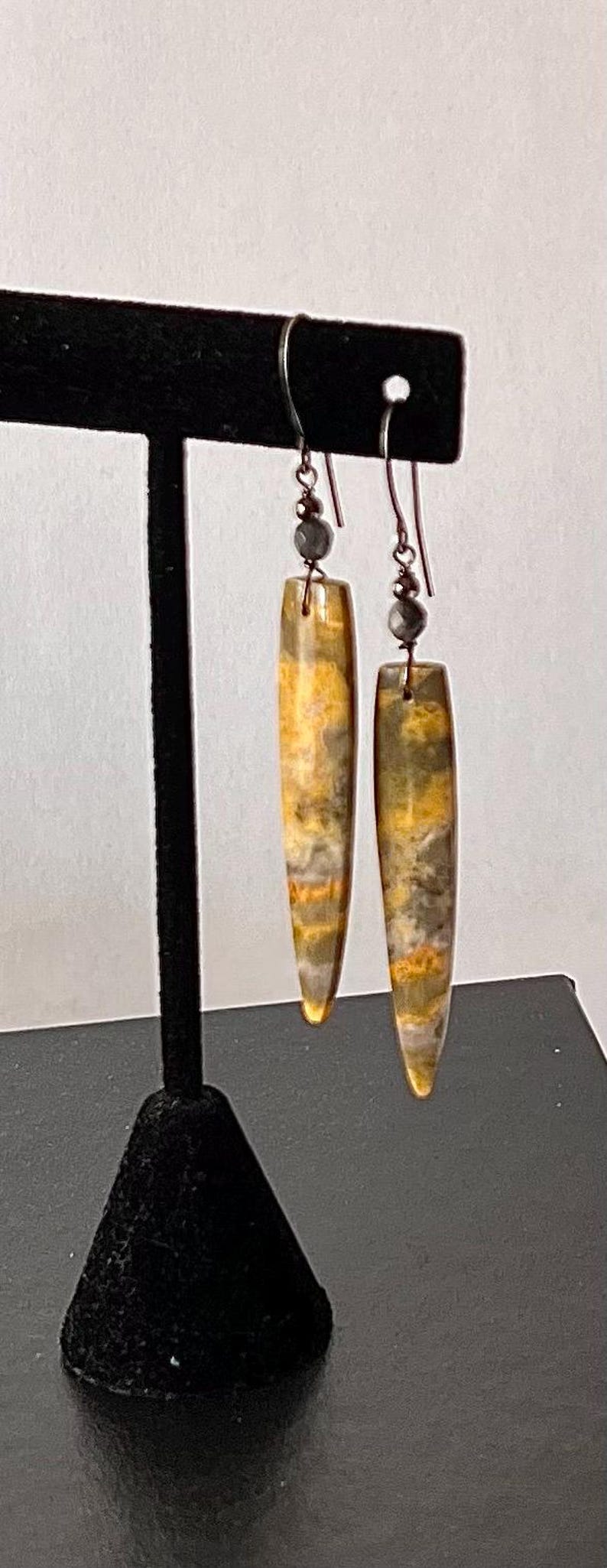 以下が含まれることがあります： 黄色、グレー、茶色の模様が入った、長方形のイヤリングのペア。 イヤリングは濃い色の金属製のフックから吊り下げられており、本体の上には小さな灰色のビーズが付いています。 イヤリングは黒いスタンドに展示されています。