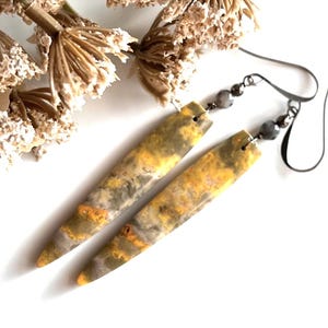 以下が含まれることがあります： 黄色、グレー、オレンジの大理石模様の、細長い長方形のイヤリングのペア。イヤリングは濃い灰色のイヤーフックと小さな灰色のビーズが付いています。背景にはドライフラワーがあります。