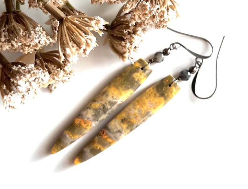 以下が含まれることがあります： 黄色、グレー、オレンジの大理石模様の、細長い長方形のイヤリングのペア。イヤリングは濃い灰色のイヤーフックと小さな灰色のビーズが付いています。背景にはドライフラワーがあります。