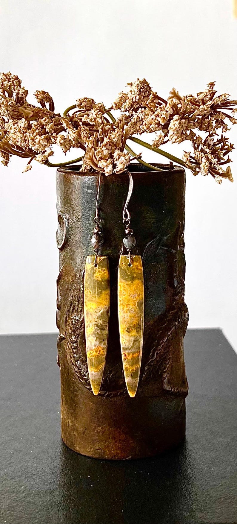 以下が含まれることがあります： 長方形の形のぶら下がりイヤリングのペア。イヤリングは、黄色と茶色の模様で、銀色のフックと小さなビーズが付いています。イヤリングは、ドライフラワーが入った暗い花瓶の前に飾られています。