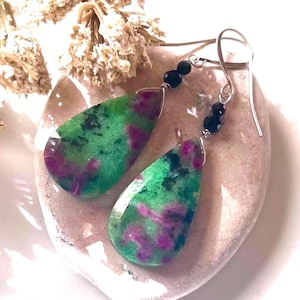 May include: A pair of teardrop-shaped earrings with green and magenta stones. The stones are faceted and have a speckled appearance. The earrings are suspended from silver-colored hooks and feature small black beads.