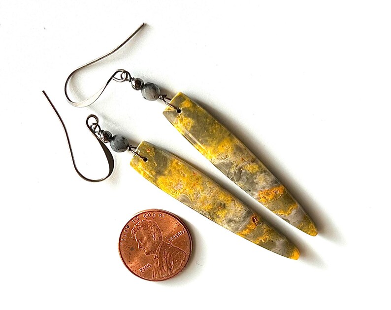 以下が含まれることがあります： 細長い黄と灰色の石のペンダントが特徴の、手作りのイヤリングのペア。イヤリングには銀色のイヤーフックと小さな灰色のビーズが付いています。サイズの目安としてペニーが含まれています。