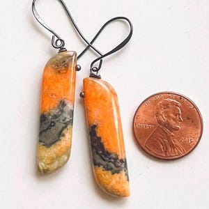 May include: A pair of orange and gray stone earrings with dark metal hooks. The earrings are rectangular with rounded edges. A penny is next to the earrings for size comparison.