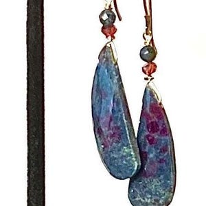 May include: Teardrop-shaped earrings with a blue and purple marbled effect. The earrings have gold-colored hooks and are accented with small red and blue beads. The earrings are displayed on a black stand.