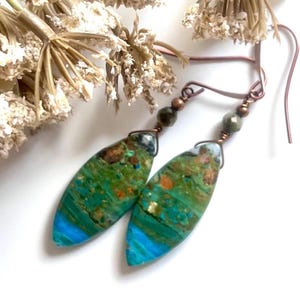 May include: A pair of handcrafted earrings featuring elongated, teardrop-shaped stones with a vibrant mix of green, blue, and brown hues. The stones are suspended from copper-colored ear wires and accented with small, dark beads. The earrings are set against a white background.