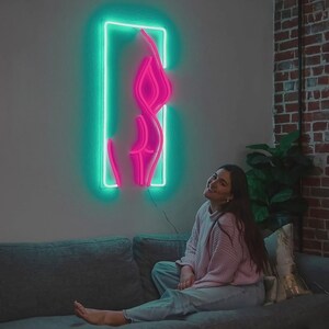 Peut inclure: Une enseigne au néon rose vif représentant une silhouette féminine, encadrée d'un rectangle vert fluo, est accrochée à un mur gris clair. Une personne est assise sur un canapé gris, regardant l'œuvre d'art. La pièce a un mur de briques et une plante.
