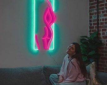 May include: A neon sign in the shape of a female form, outlined in bright pink, is encased in a rectangular frame of green neon. The sign is mounted on a light gray wall. A person is sitting on a gray couch, looking up at the sign.