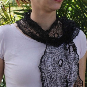 May include: A woman wearing a black lace scarf with a delicate, open-weave pattern. The scarf is draped around her neck and falls down her chest. She is wearing a plain white shirt.