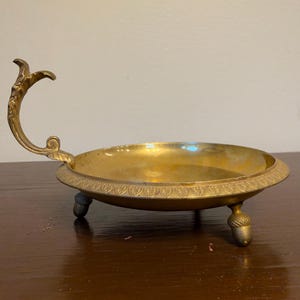May include: A round brass dish featuring a curved handle with ornate detailing and a decorative rim pattern. The dish is supported by three small feet shaped like acorns, providing a stable base for this metallic serving piece.