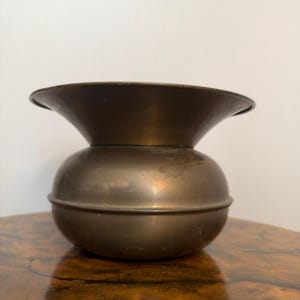 May include: A metallic, bronze-toned vessel featuring a rounded, bulbous base and a wide, flared rim. The object has a smooth, uniform surface with a subtle sheen, characterized by a distinct horizontal seam separating the lower body from the neck.