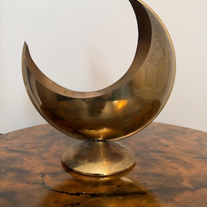 May include: A crescent moon-shaped brass bowl with a smooth, polished finish and a circular pedestal base. The metallic object features a curved, open top design and a warm, golden-toned surface with subtle signs of natural aging.