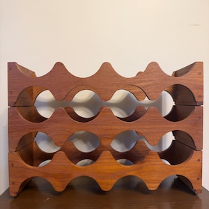 May include: A three-tiered wooden wine rack featuring a repeating wave-like pattern with circular cutouts designed to hold bottles. The structure is constructed from medium-toned wood with visible grain and is held together by small metal screws.