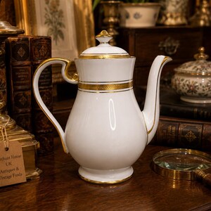 Pode incluir: Uma cafeteira de porcelana branca com alça curva, bico cônico e tampa com remate pontiagudo. A cafeteira é decorada com faixas horizontais de borda dourada ao redor da tampa, do pescoço e da base, com detalhes dourados na alça.