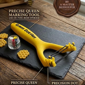 Puede incluir: Una herramienta de marcado amarilla "Queen Dot-Her" para apicultura, con bandas negras y almacenamiento de bandas. La herramienta está sobre una superficie de pizarra negra, con un tarro de puntos de colores y decoraciones en forma de panal.