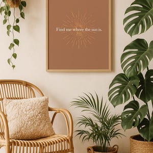 Peut inclure: Une affiche encadrée avec le texte "Find me where the sun is" et un motif de soleil. L'affiche est accrochée au-dessus d'une chaise en rotin avec un coussin moelleux. Des plantes en pot et un panier tressé sont également visibles.