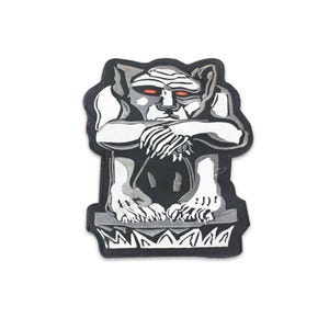 Könnte beinhalten: Ein Aufnäher mit einer stilisierten Wasserspeier-Figur in Grau- und Weißtönen mit roten Augen. Die Wasserspeier-Figur ist mit verschränkten Armen dargestellt und sitzt auf einem dekorativen Sockel. Der Aufnäher hat einen schwarzen Rand.