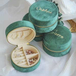以下が含まれることがあります： 金色のジッパーが付いた緑色のベルベット製のジュエリーボックスが3つあります。ボックスには「Daphne」、「Sierra」、そしてもう1つの名前が刻印されていますが、見えません。中央のボックスは開いており、指輪、イヤリング、ネックレスを入れるための仕切りがあります。