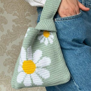 May include: A light green crocheted bag with a white daisy pattern. The bag has a knotted handle and is made with a soft, textured yarn.