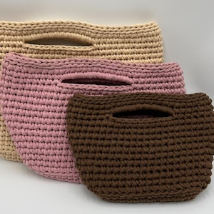 May include: Three handmade crochet bags in varying sizes and colors. The largest bag is a light beige, the middle bag is pink, and the smallest is brown. Each bag has a handle opening at the top, and the bags are made of a thick, textured yarn.
