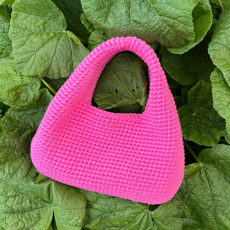 May include: A bright pink, crocheted handbag with a rounded shape and a single handle. The bag is set against a backdrop of large, green leaves. The texture of the crochet is visible, adding to the handmade aesthetic.