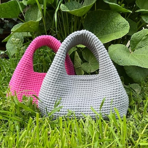 May include: Two handmade crochet bags. One is a vibrant pink, the other a soft gray. Both have a rounded shape with a sturdy handle. The bags are set against a backdrop of green foliage and grass, highlighting their texture and color.