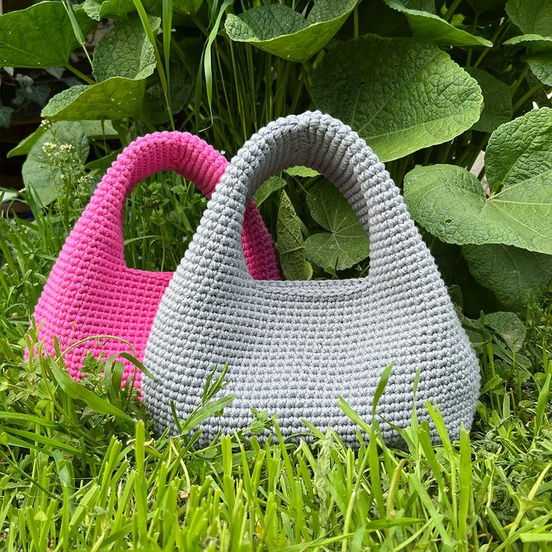 May include: Two handmade crochet bags. One is a vibrant pink, the other a soft gray. Both have a rounded shape with a sturdy handle. The bags are set against a backdrop of green foliage and grass, highlighting their texture and color.