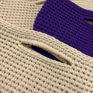 May include: Close-up of three crocheted bags in cream and purple. The cream-colored bag has a handle with a hole. The purple bag is in the background. The bags are made of thick yarn and have a textured appearance.